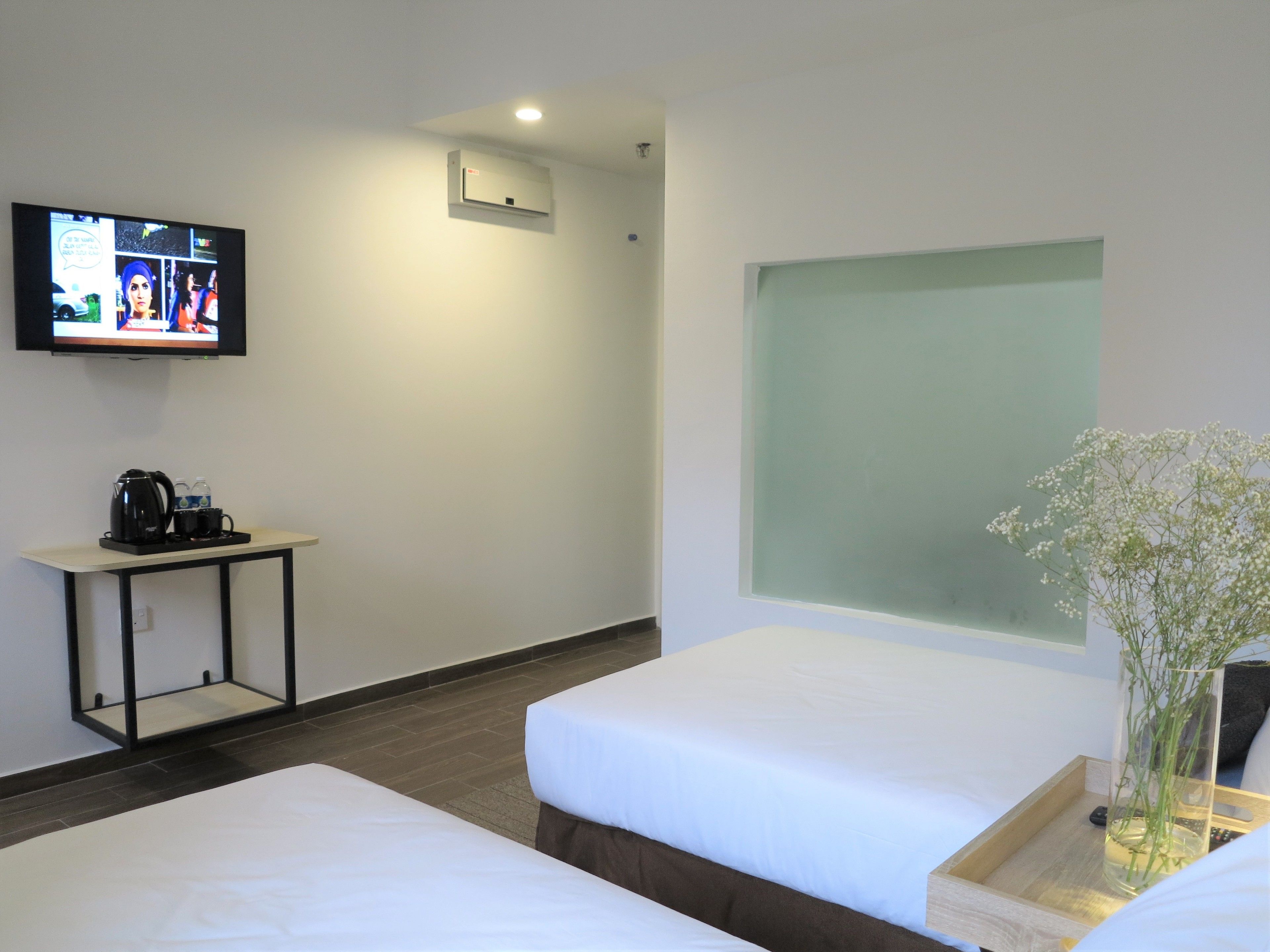 Habitación de hotel con dos camas, un televisor en la pared, una mesita con cafetera y un jarrón con flores.