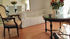 A living room with a white sofa, a wooden chair, a glass table with a vase of flowers, and a lamp.