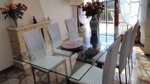 A dining area with a glass table and patterned chairs, a vase with flowers, and a view through large windows.