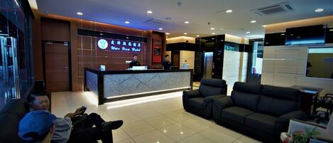 A hotel reception area with a modern design, including a reception desk, seating area, and a large screen displaying the hotel name.