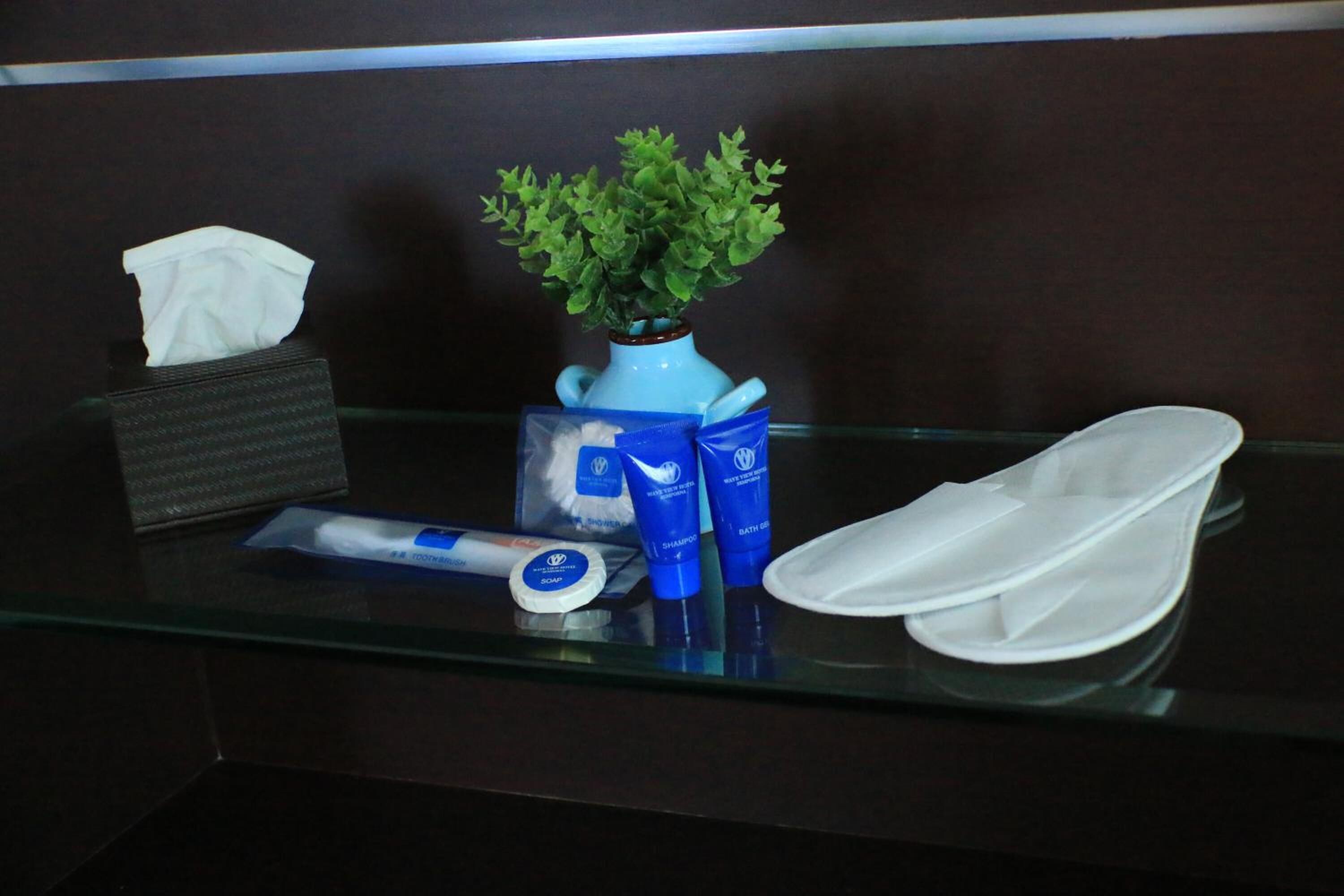A hotel reception area with a glass counter. On the counter, there is a tissue box, a blue vase with a green plant, a blue shampoo bottle, a white bath cap, and a white face mask.