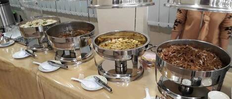 Een buffetopstelling met verschillende roestvrijstalen warmhoudbakken vol met diverse gerechten, inclusief bestek en borden.