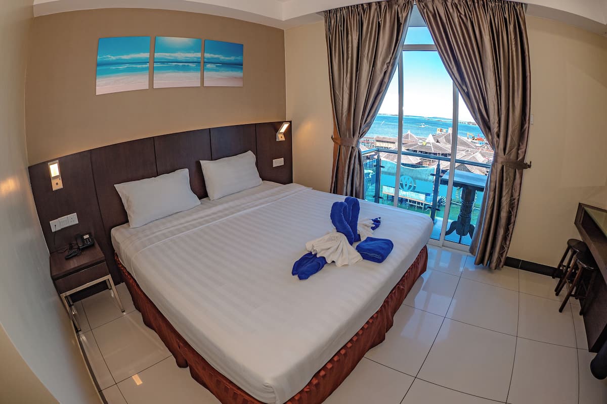 A hotel room with a large bed, a nightstand, a desk with a chair, and a view of the sea through the window.