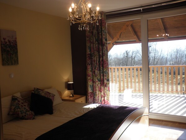 A bedroom with a bed, a nightstand, a lamp, a chandelier, and a view of a wooden deck through a sliding glass door.