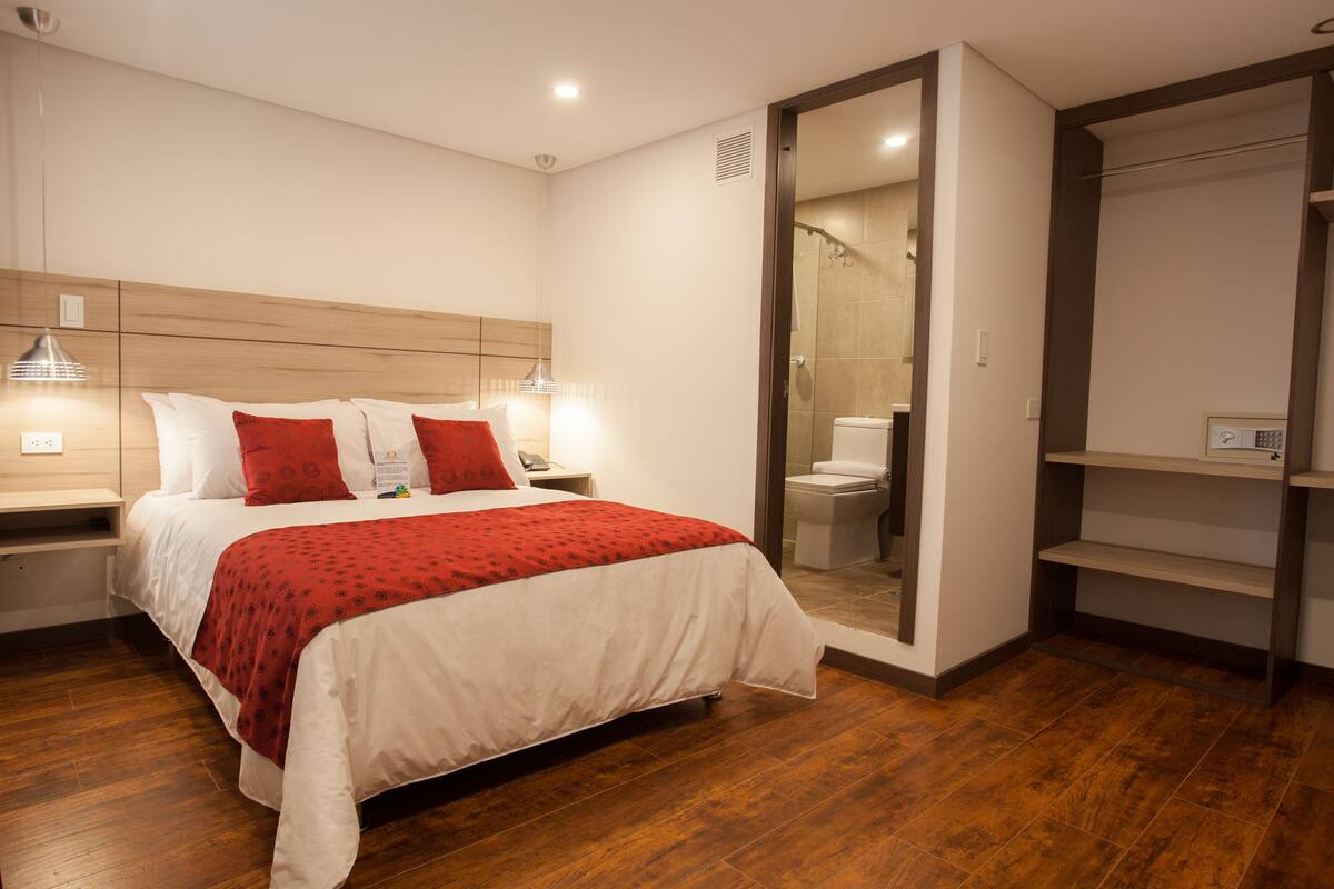 A modern hotel room with a wooden floor, a bed with white bedding and red accents, a bedside table with a lamp, and a large mirror.
