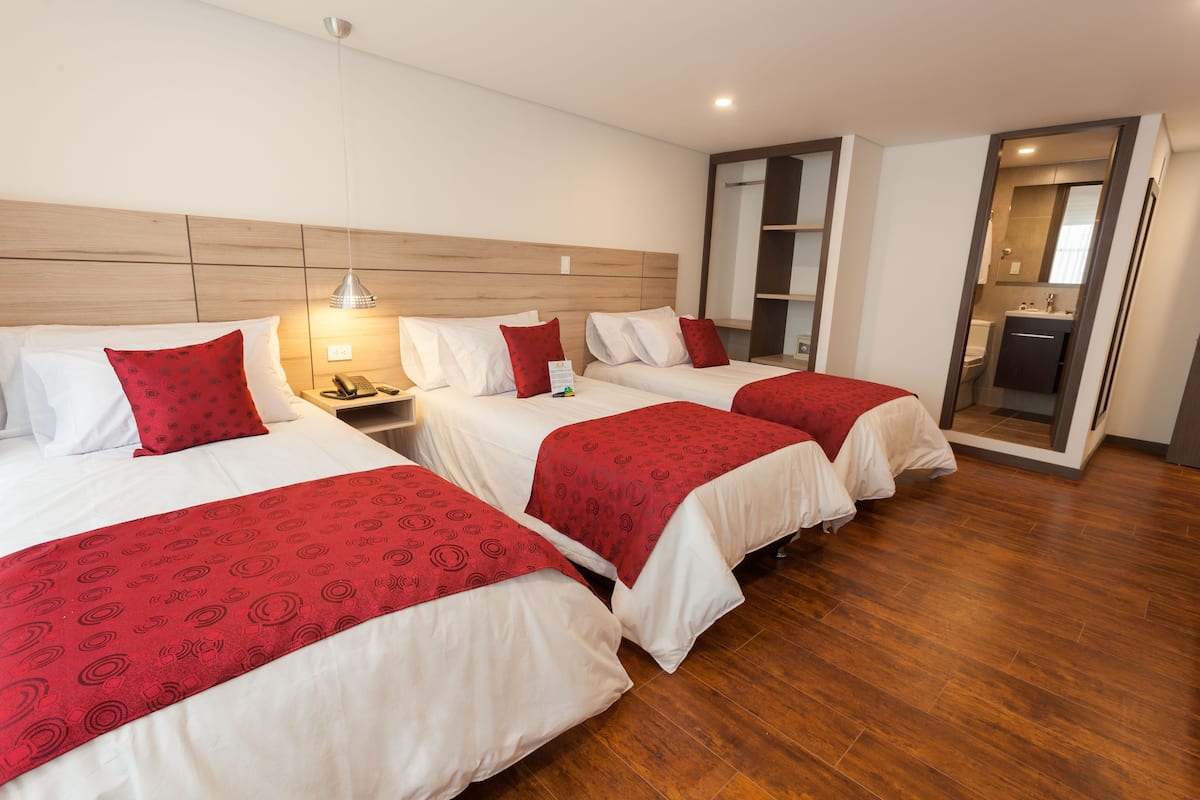 A hotel room with three beds, each with red and white bedding, a wooden headboard, and a bedside table with a lamp.