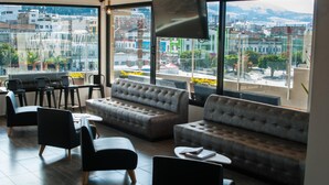 Un moderno salón con sofás de cuero y sillas negras, ventanas amplias con vistas a la ciudad y un televisor de pantalla plana montado en la pared.