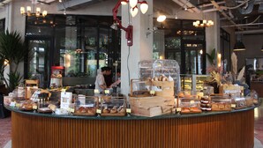 A bakery counter with various baked goods displayed in clear containers, a cashier, and a modern interior with exposed pipes and natural light. - Hotel Monopoli (Jakarta)