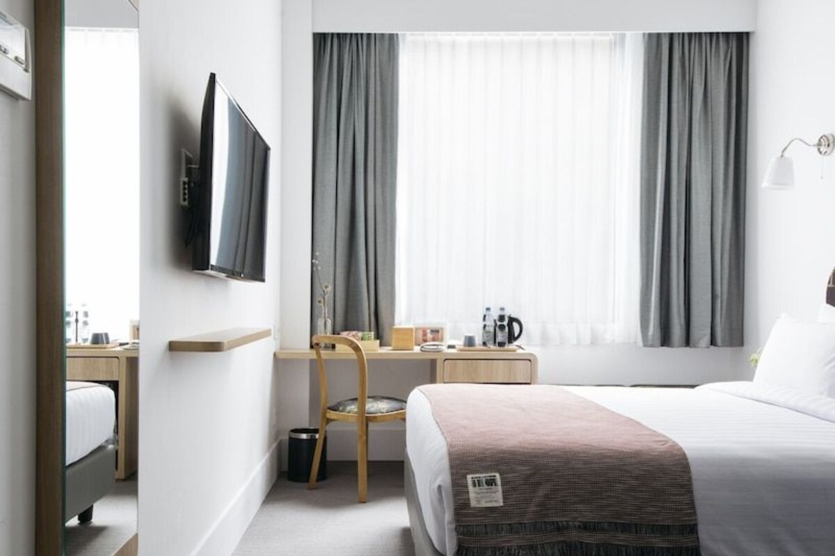 A modern hotel room with a bed, a desk, a chair, a TV, and a large mirror.