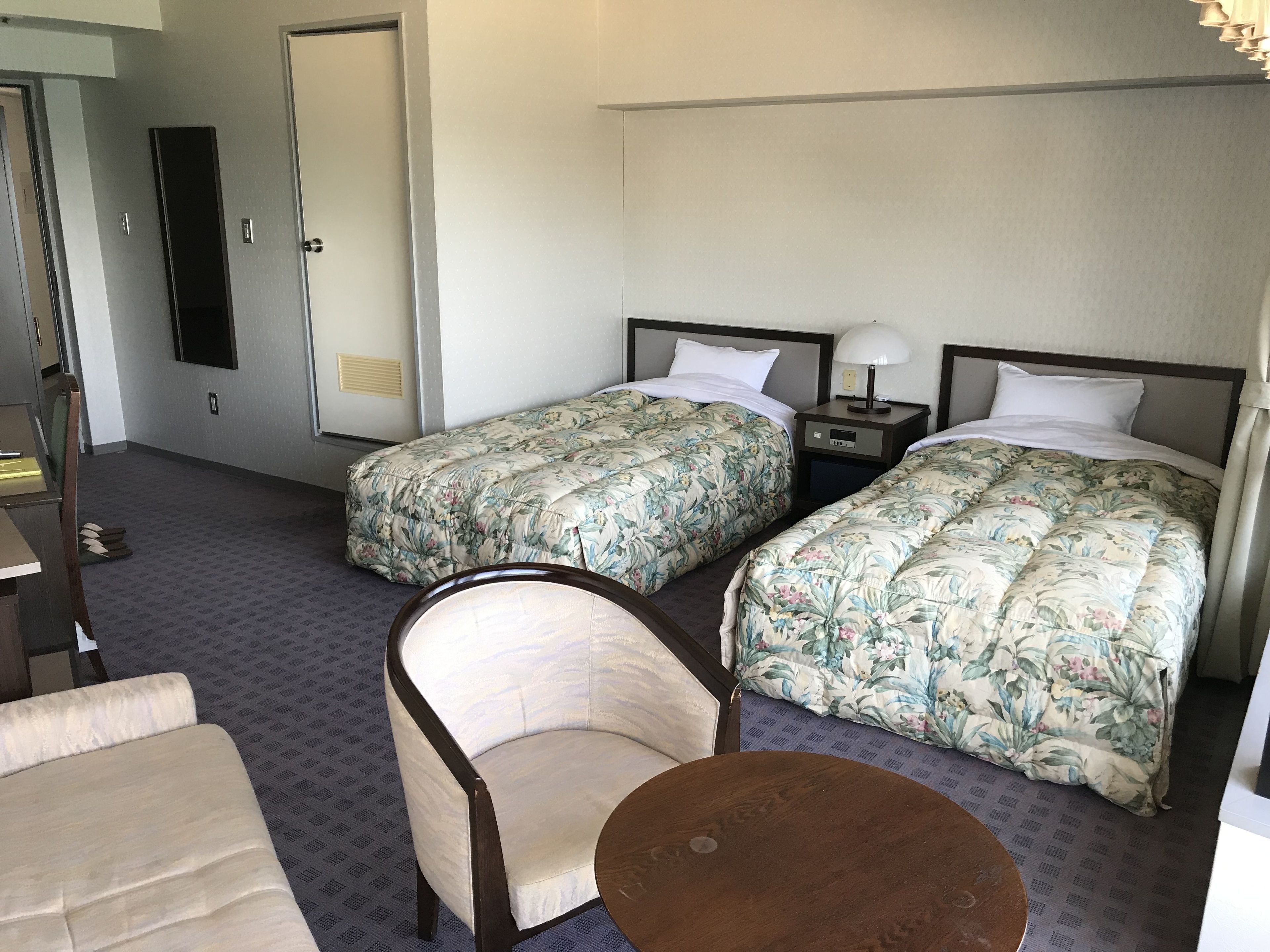 Ein Hotelzimmer mit zwei Betten, einem Sessel, einem kleinen Tisch und einem Schreibtisch mit Lampe.