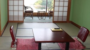 Une pièce avec un sol recouvert de tatamis, une cloison coulissante de type shoji, une table en bois, deux chaises et une vue sur une zone boisée.