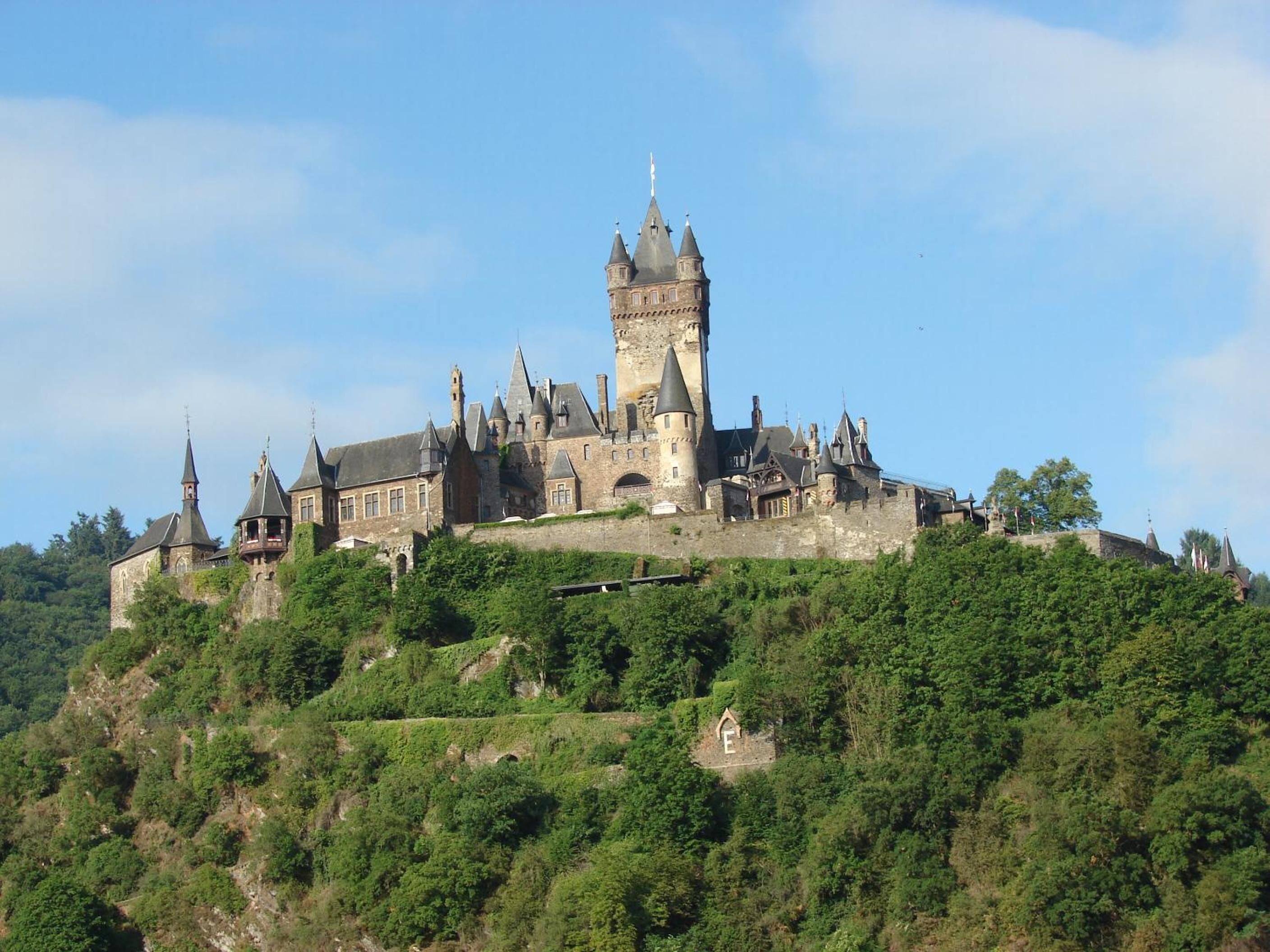 A historic castle with multiple towers and a prominent central tower, surrounded by lush greenery and perched on a hill.