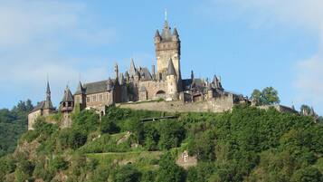 A historic castle with multiple towers and a prominent central tower, surrounded by lush greenery and perched on a hill.