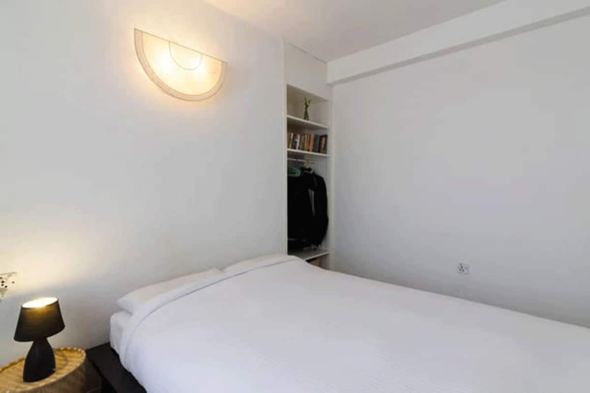 Un dormitorio pequeño y ordenado con cama, lámpara de noche, luz en la pared y estantería empotrada.