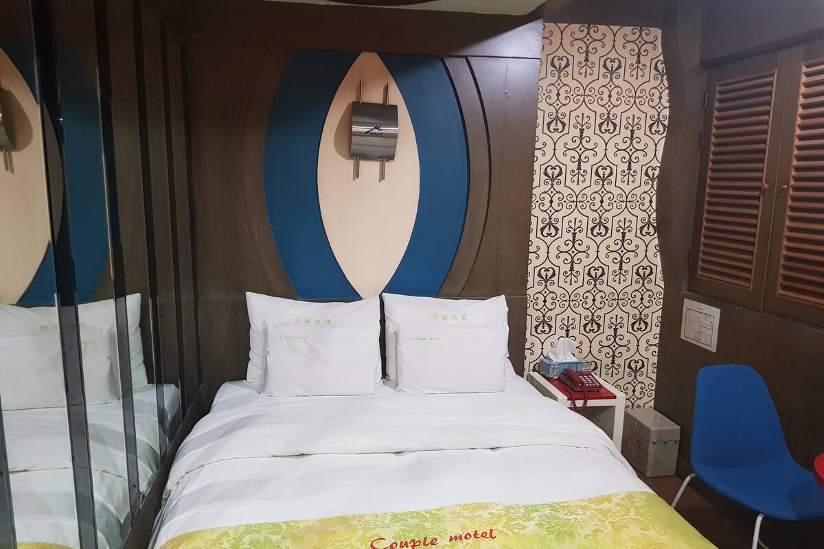 A hotel room with a bed, a bedside table with a telephone and a box, a blue chair, and a patterned wall.