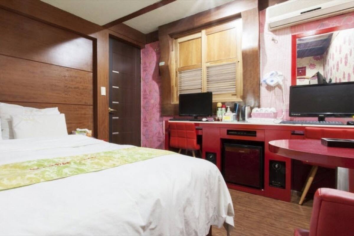 A hotel room with a bed, a desk with two monitors, and a small kitchenette.