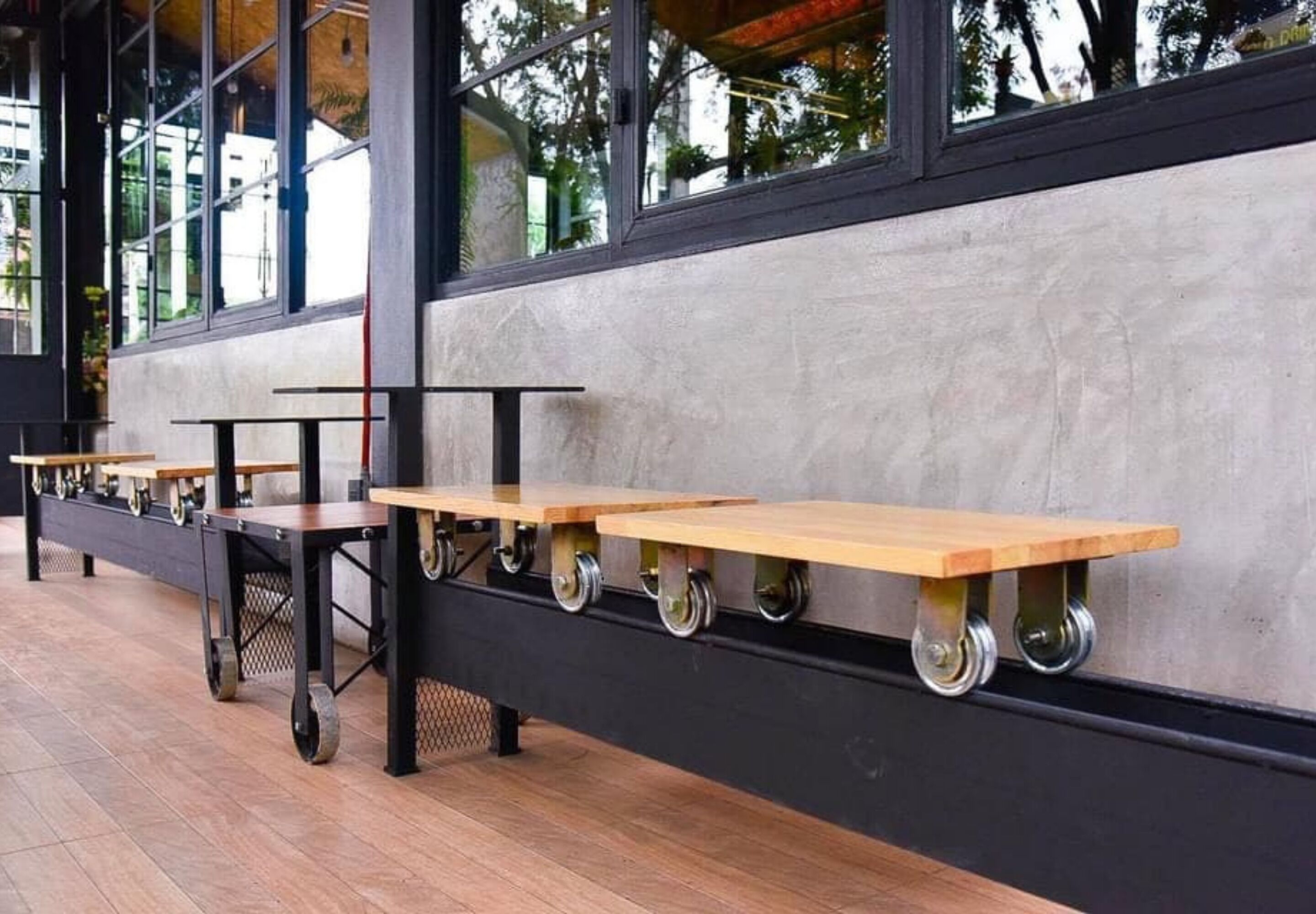 A row of wheeled tables with wooden tops, mounted on a wall with a black frame, in a covered outdoor area with a tiled floor.