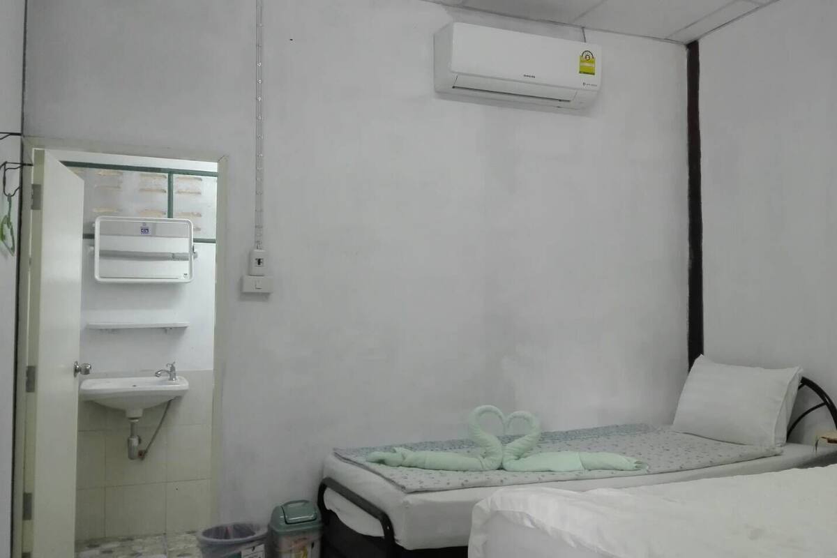A small, clean room with a bed, a bedside table with bottles, a wall-mounted air conditioner, and a bathroom with a sink and towel rack.
