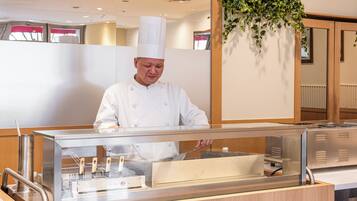 Um chef com uniforme branco atrás de um balcão de aço inoxidável com uma barreira de vidro, em um espaço bem iluminado com painéis de madeira e uma planta pendente.