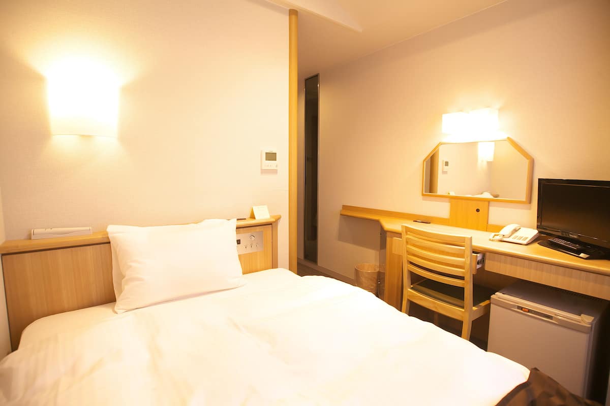 A hotel room with a bed, a desk with a chair, a desk lamp, a television, and a small refrigerator.