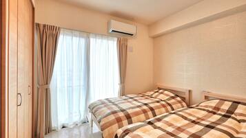 A bedroom with two beds, a window with curtains, an air conditioning unit, and a wooden wardrobe.