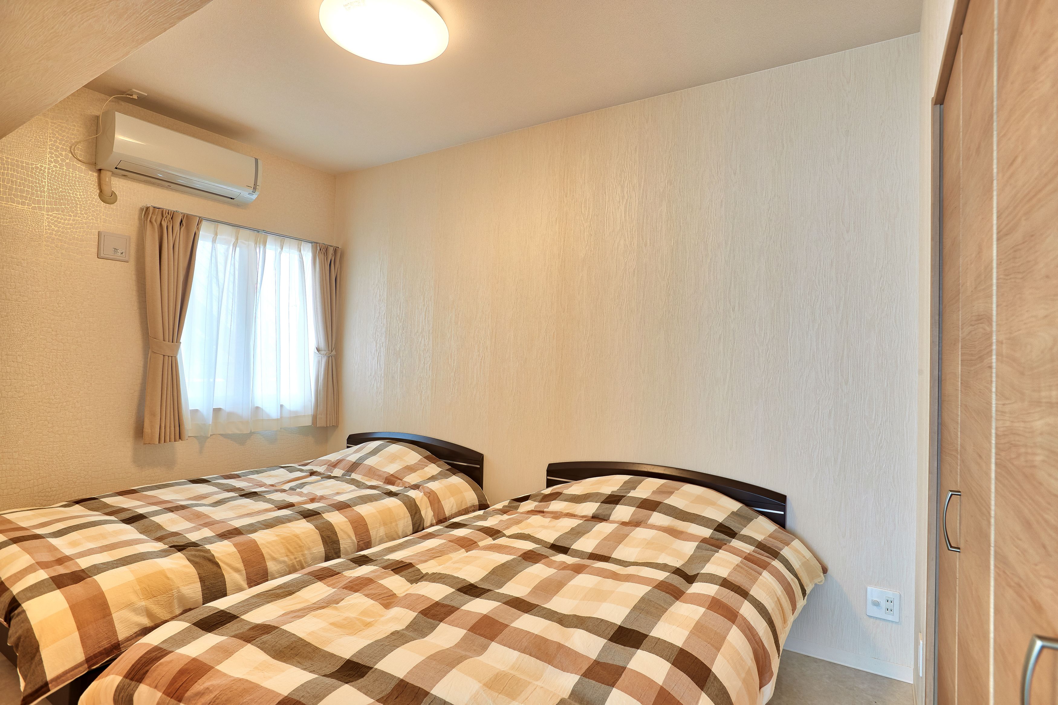 A room with two beds, a window with curtains, an air conditioning unit, and a wooden door.