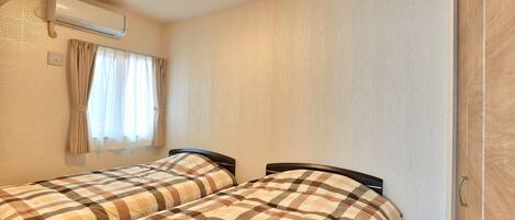 A room with two beds, a window with curtains, an air conditioning unit, and a wooden door.
