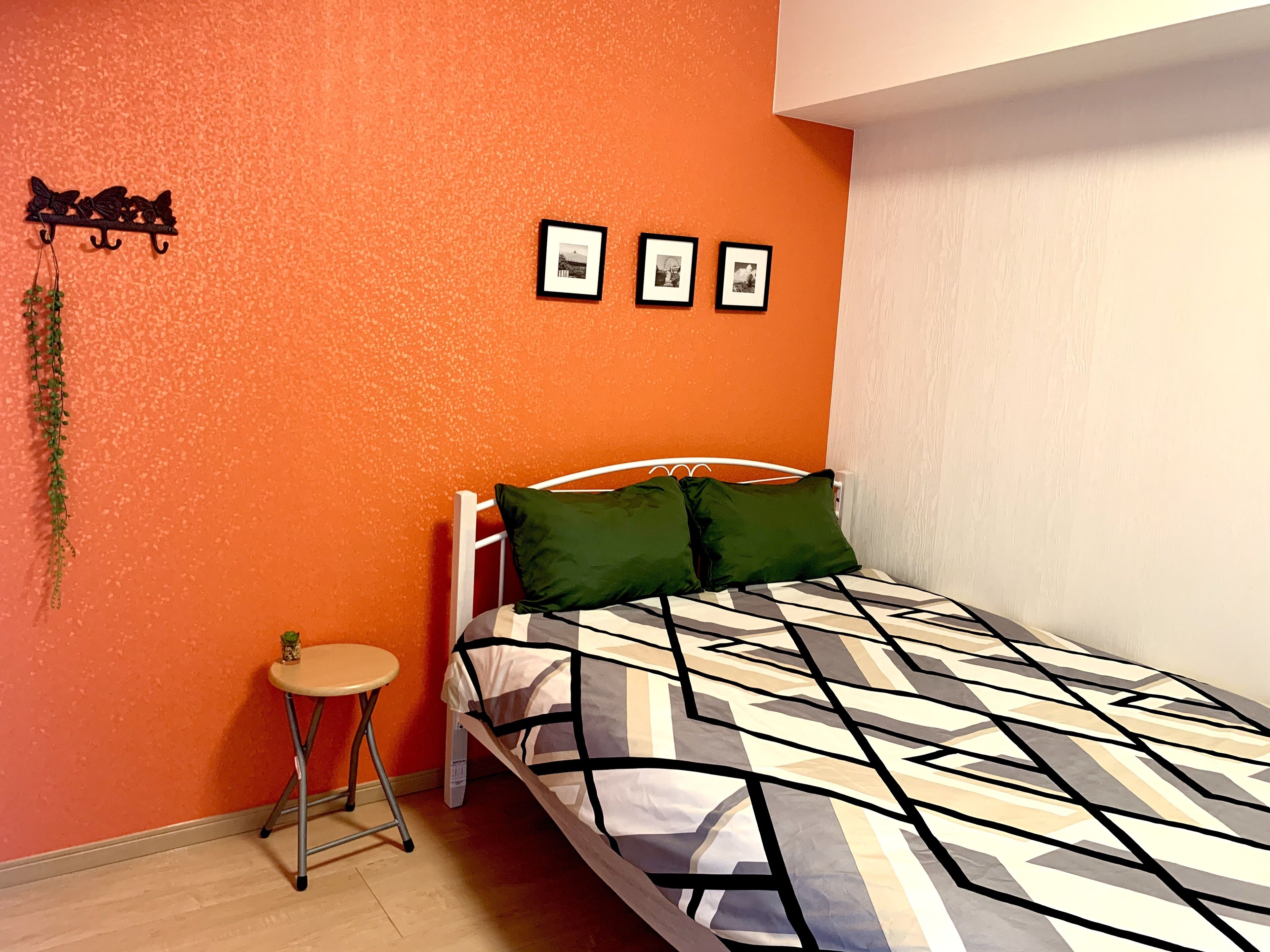 Un dormitorio con una pared naranja, una cama con colcha de estampado geométrico, dos almohadas verdes, una mesita redonda con una planta y tres cuadros en la pared.