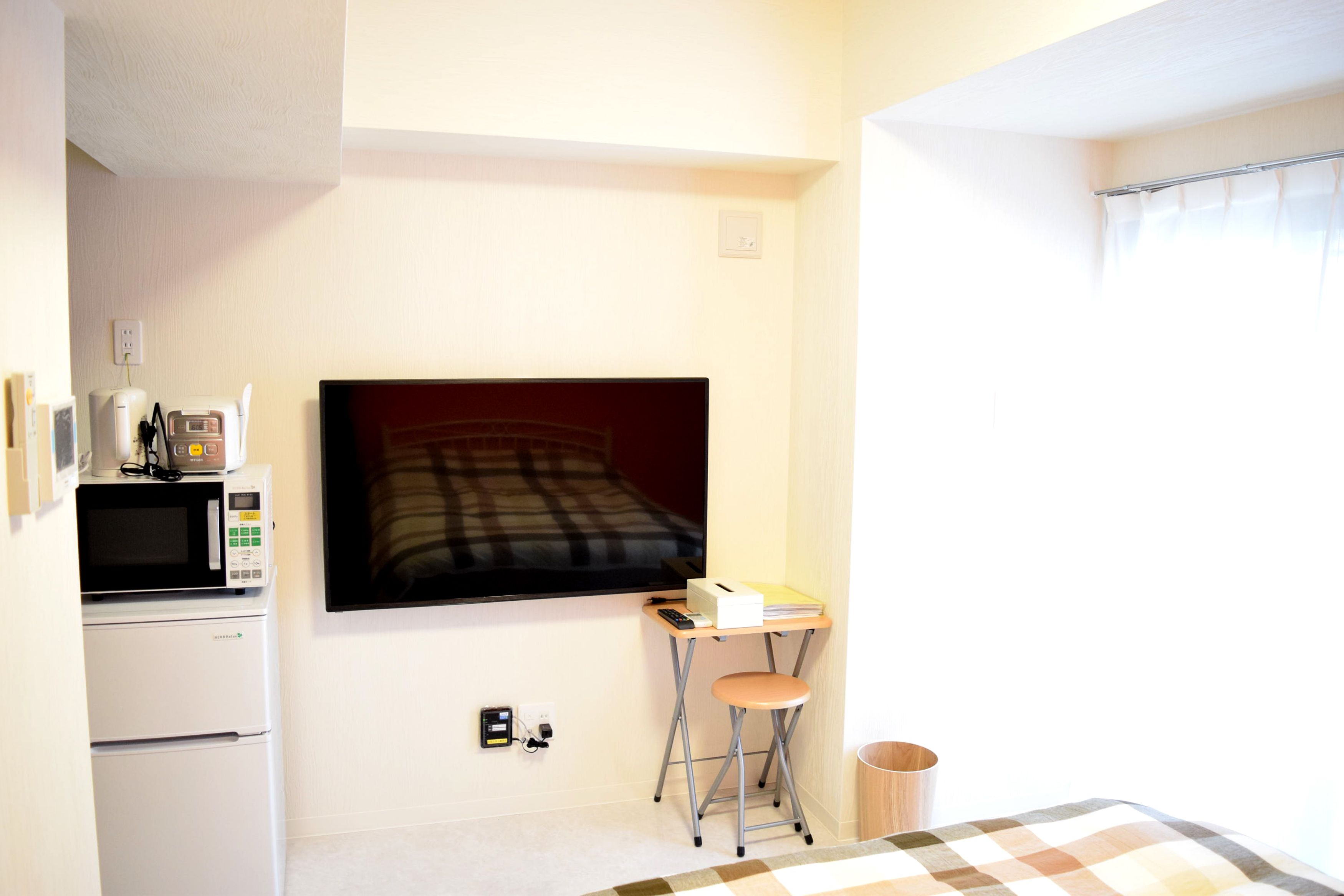 A small, well-lit room with a bed, a microwave, a refrigerator, a desk with a chair, and a window with curtains.