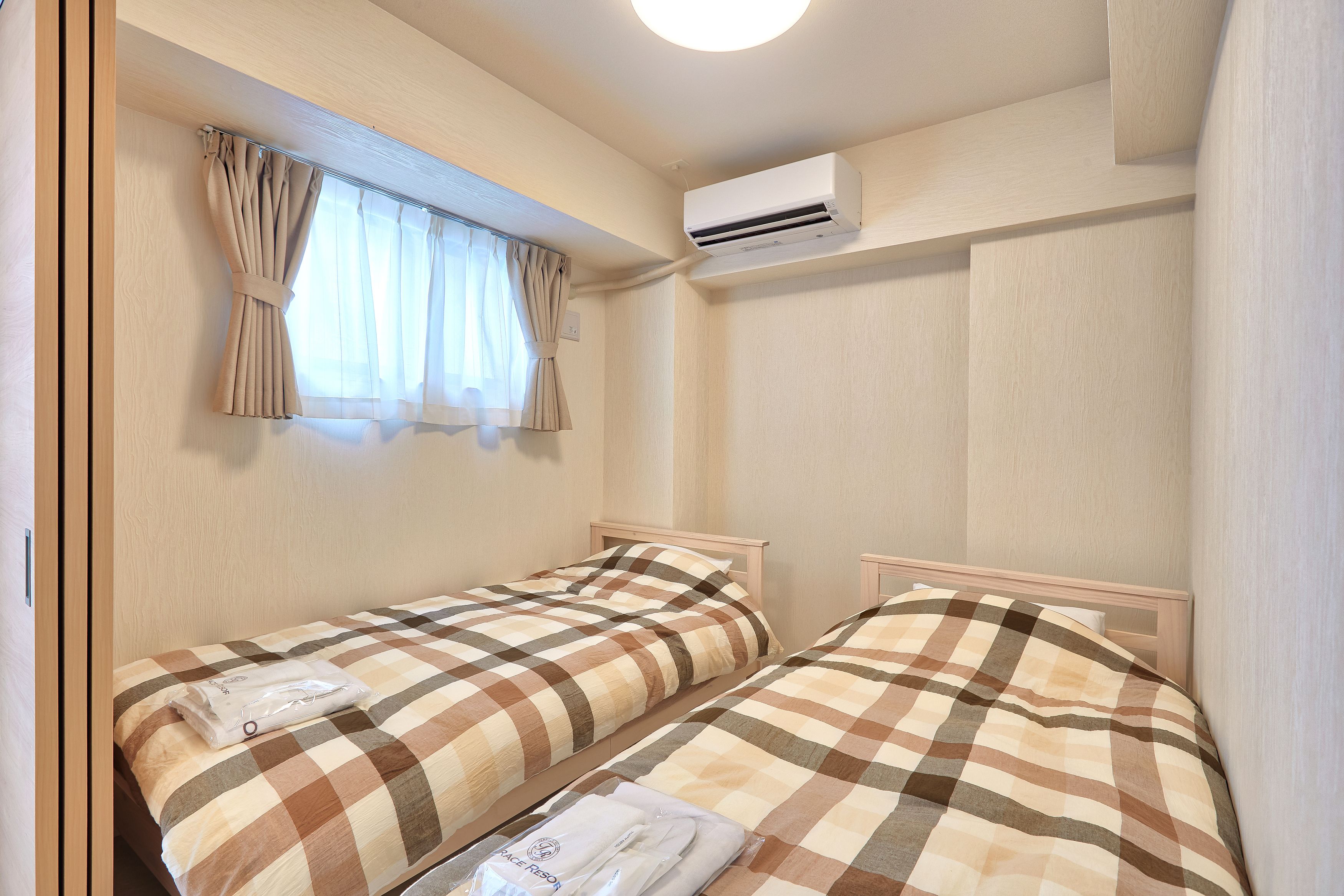 A small, neatly arranged bedroom with two beds, a window with curtains, an air conditioning unit, and a towel on one bed.
