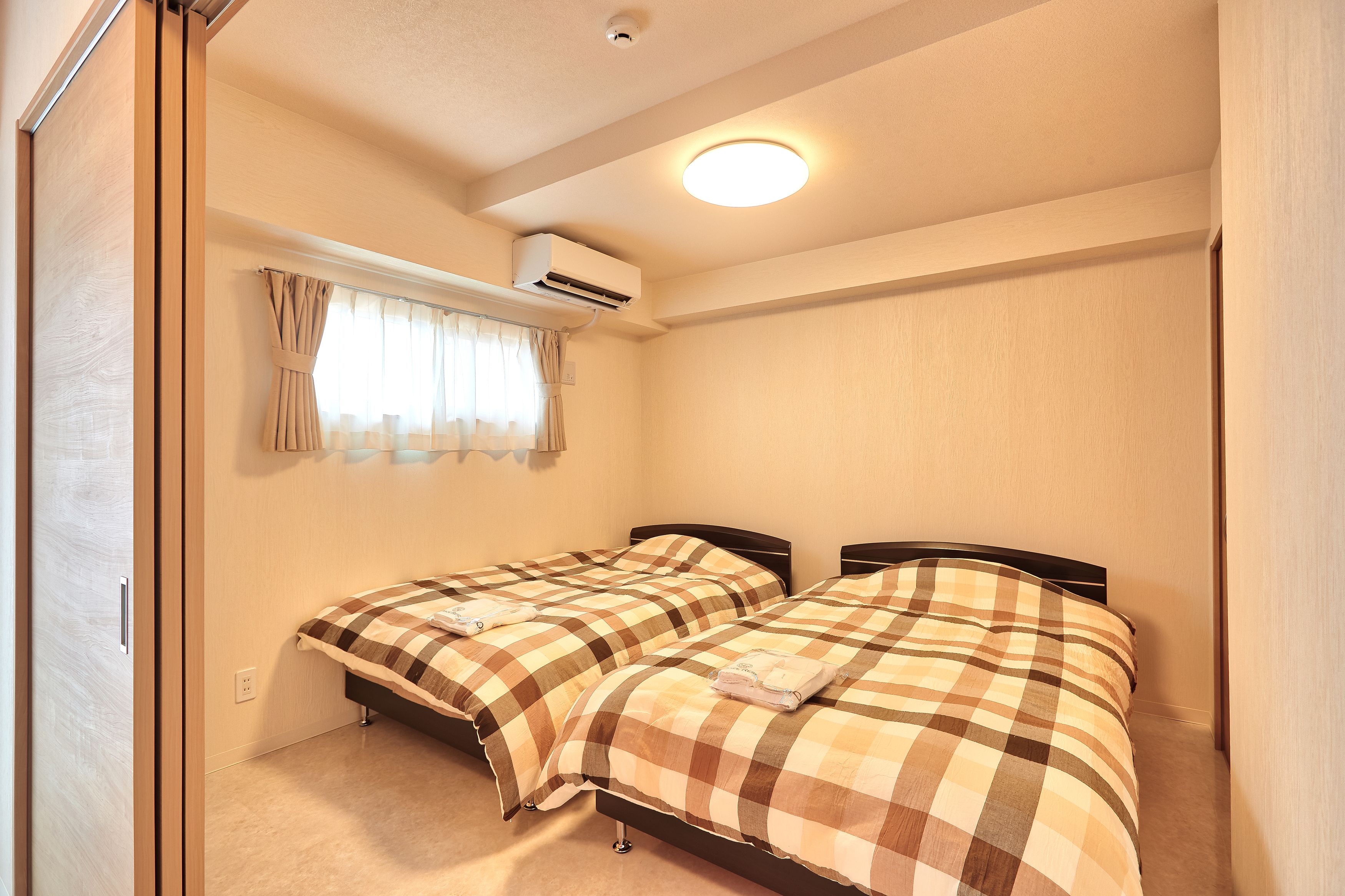 Un dormitorio con dos camas, un equipo de aire acondicionado, una ventana con cortinas y una lámpara en el techo.