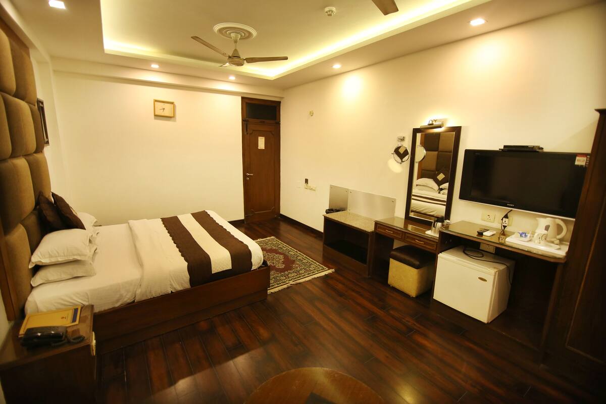Habitación de hotel con cama, escritorio con televisor, refrigerador pequeño y ventilador de techo.