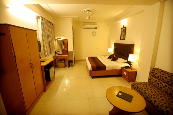 A hotel room with a bed, a sofa, a desk, a wardrobe, a mirror, a clock, and an air conditioning unit.