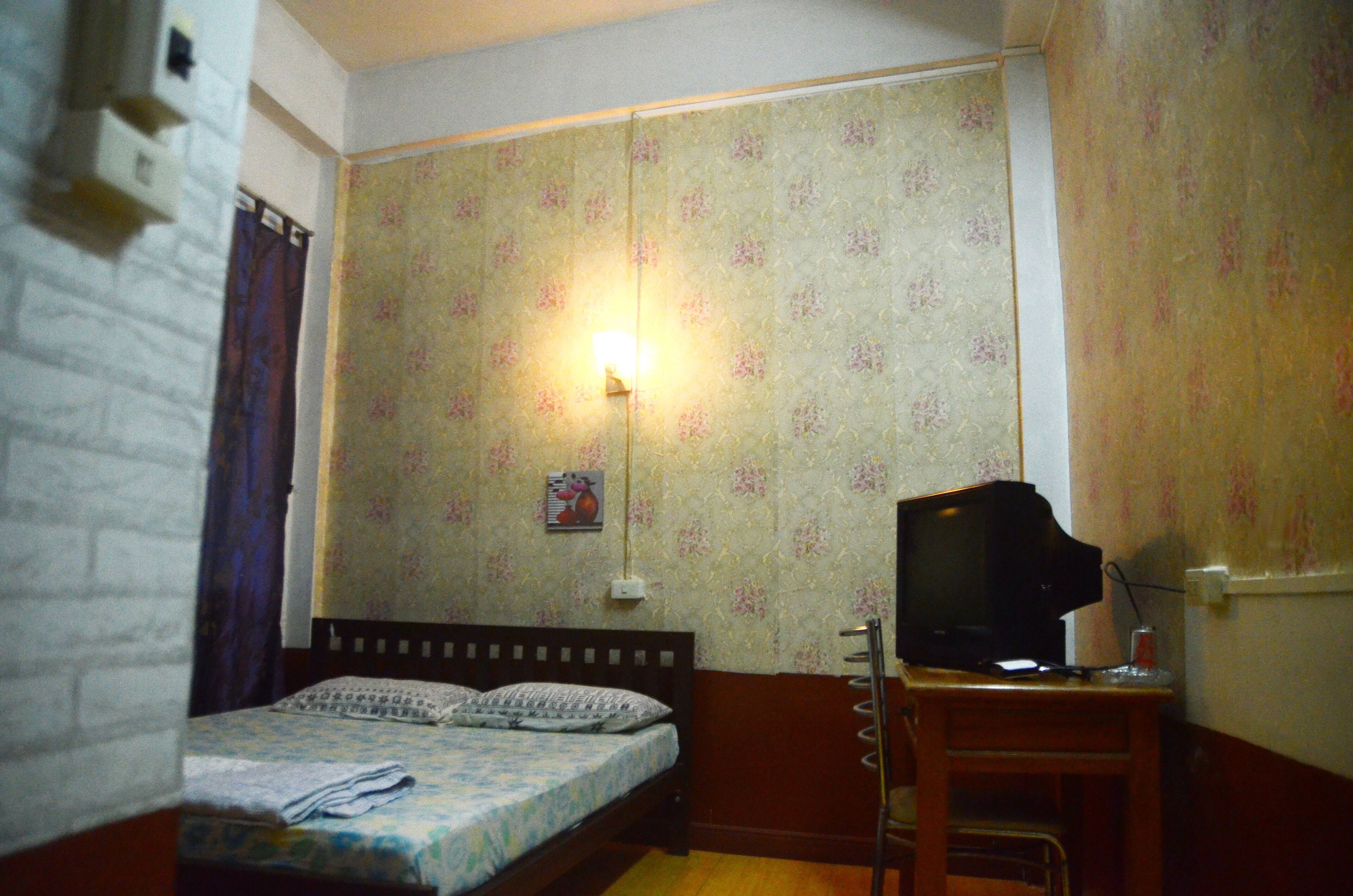 Una habitación pequeña con una cama, un escritorio con televisor y una lámpara fijada a la pared.