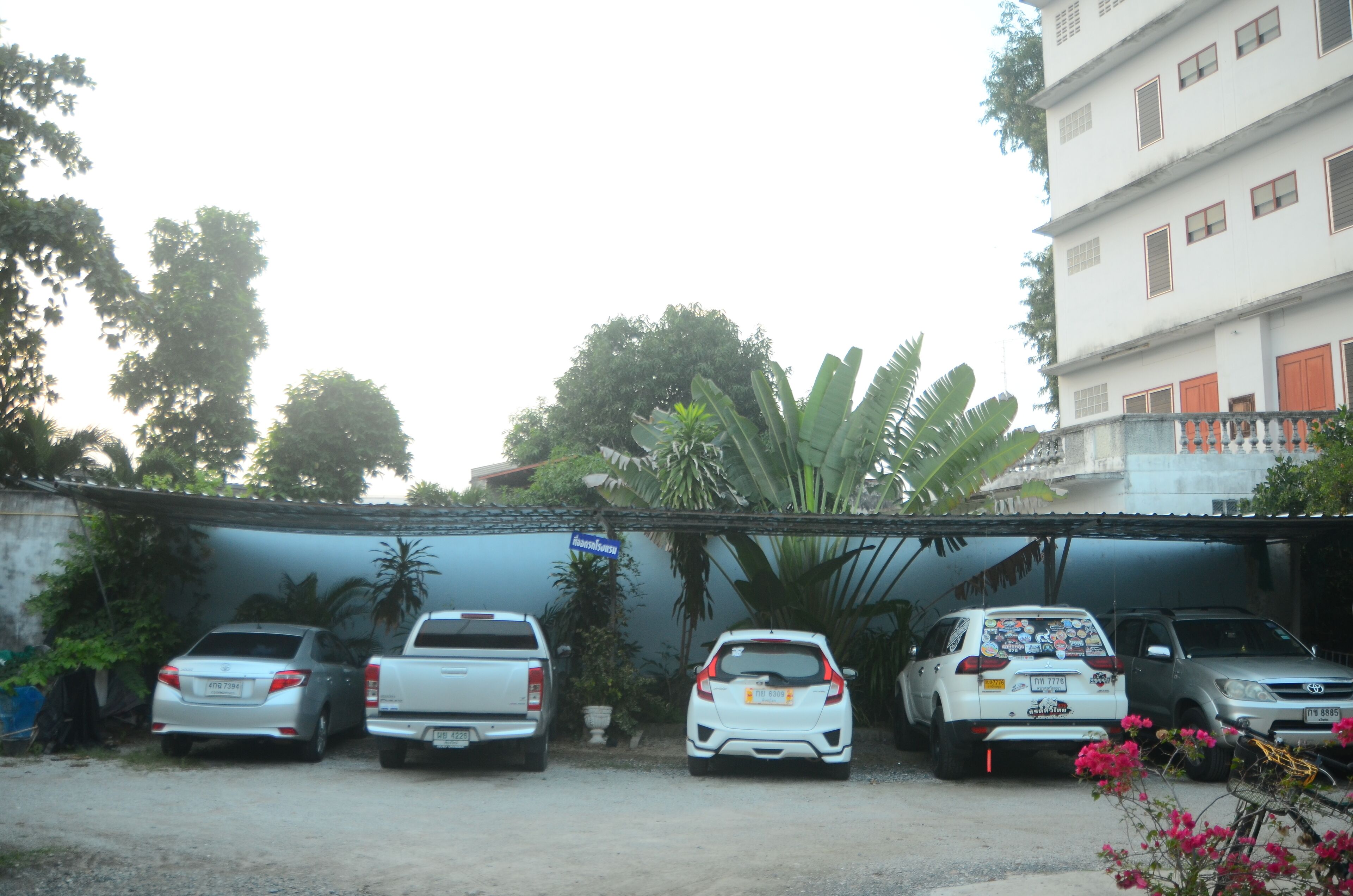 A parking area with several vehicles, including a white pickup truck, a white compact car, and a silver SUV. There are trees and a building with multiple windows in the background.
