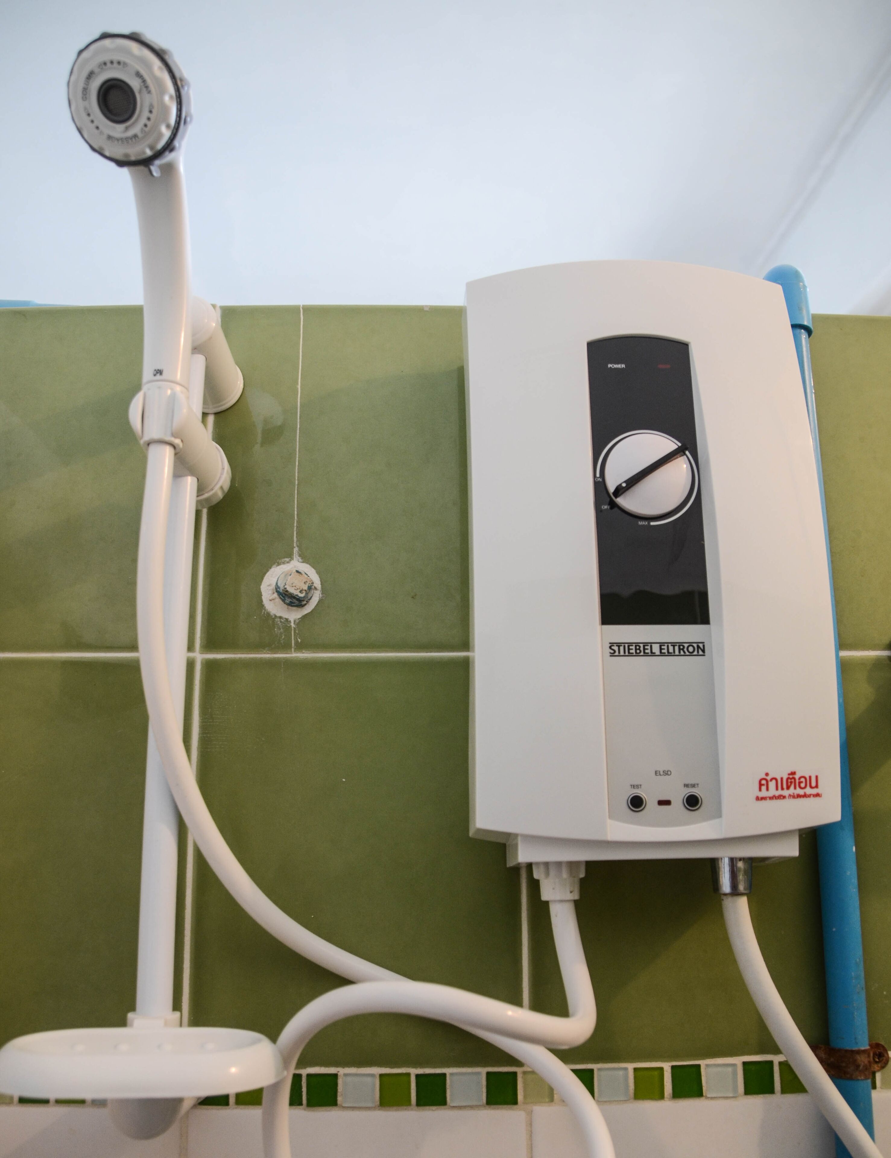 A water heater with a control panel, connected to a showerhead and pipes, against a green tiled wall.