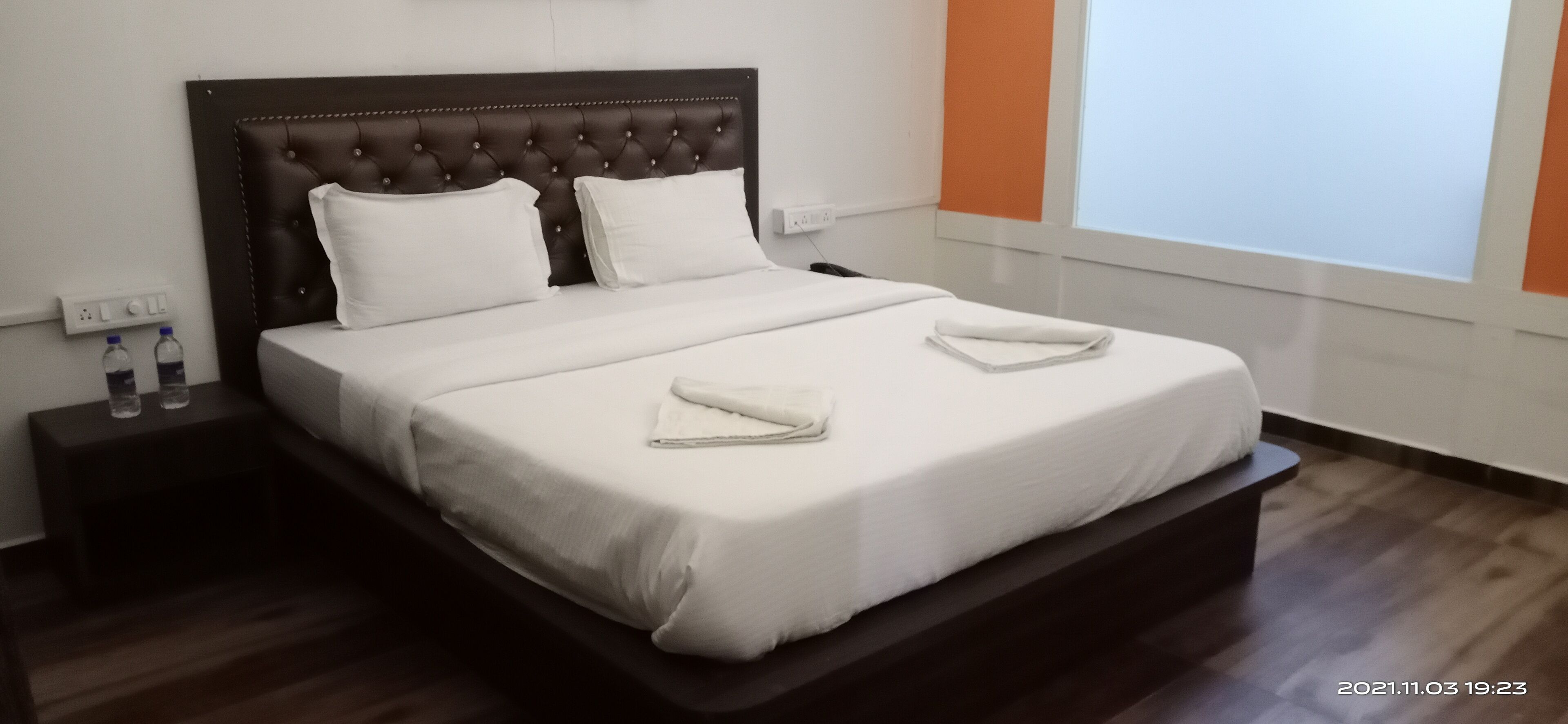 A neatly made bed with white linens and two folded towels, a dark headboard, and a nightstand with two water bottles.