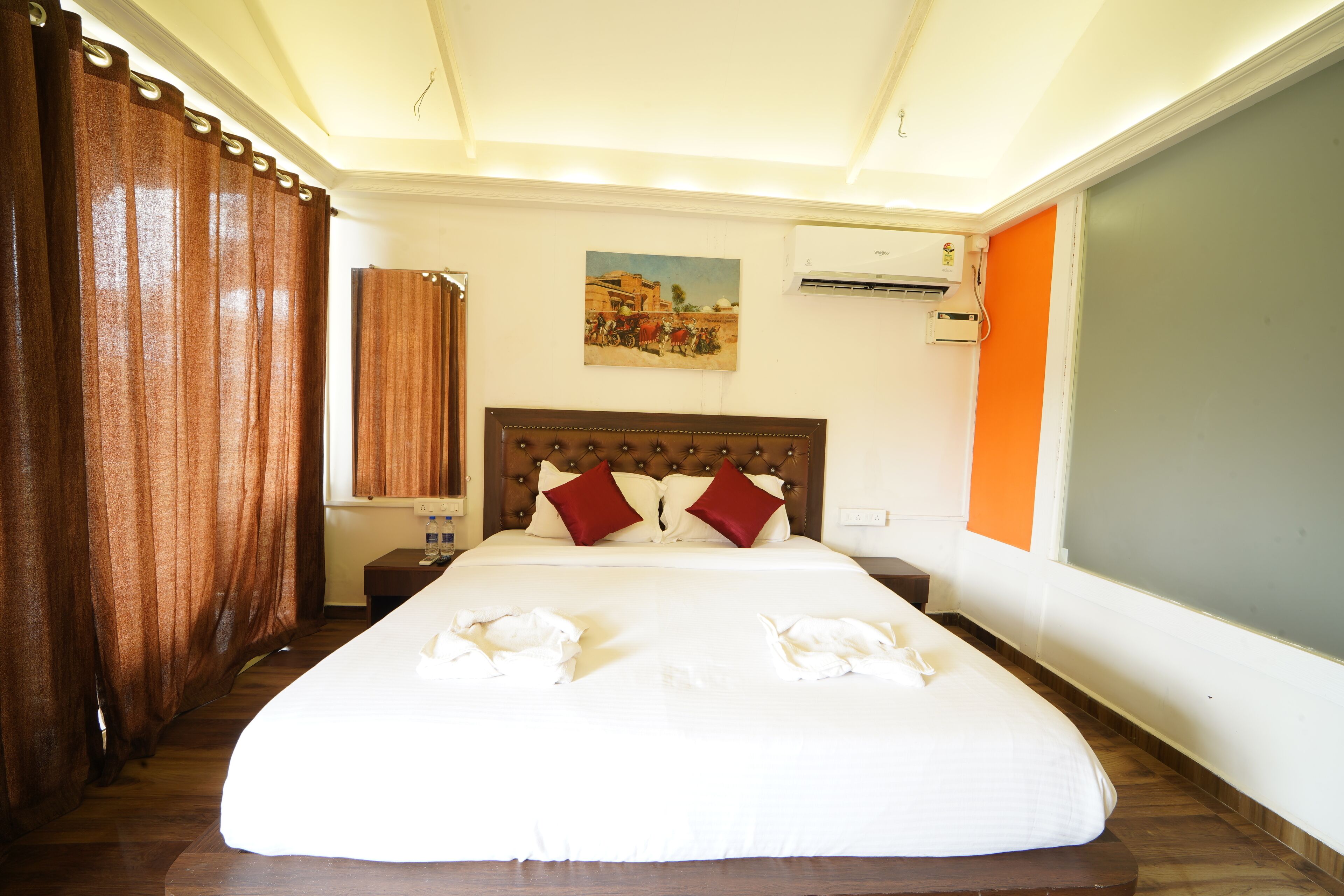 A hotel room with a large bed, a wooden headboard, two red pillows, a painting on the wall, and an air conditioning unit.