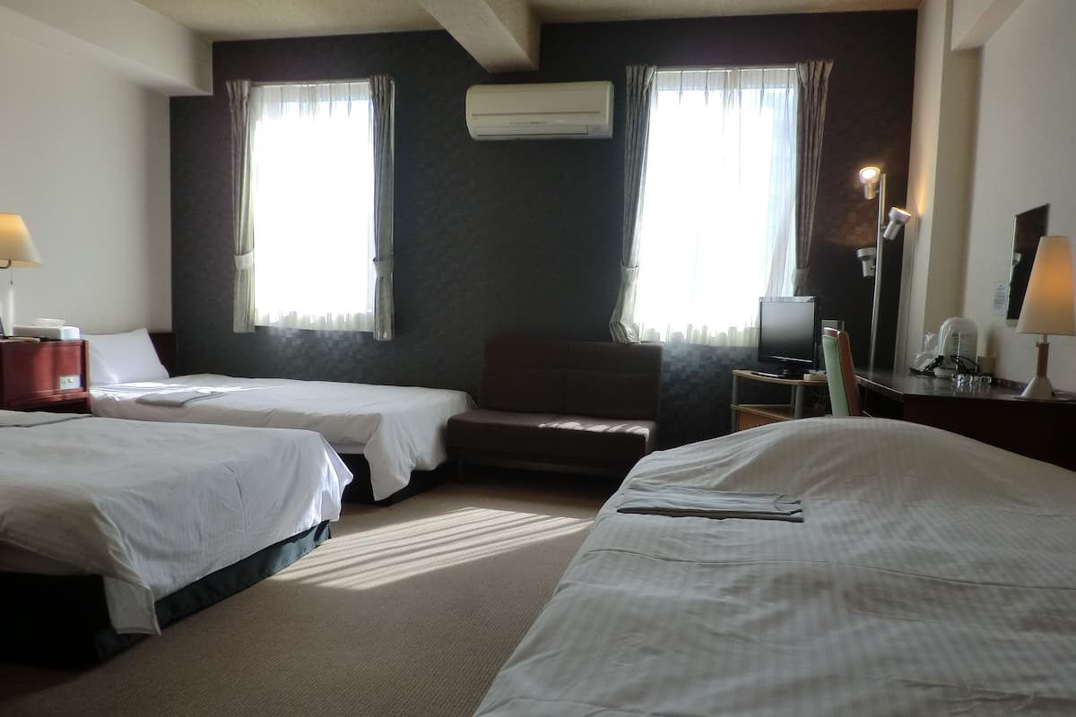 A hotel room with two beds, a sofa, a desk with a lamp, and an air conditioning unit.