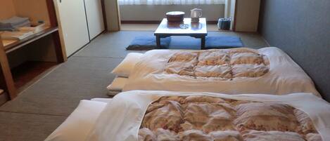A hotel room with two beds, a small table with a kettle and cups, and a view of the outside through a window.