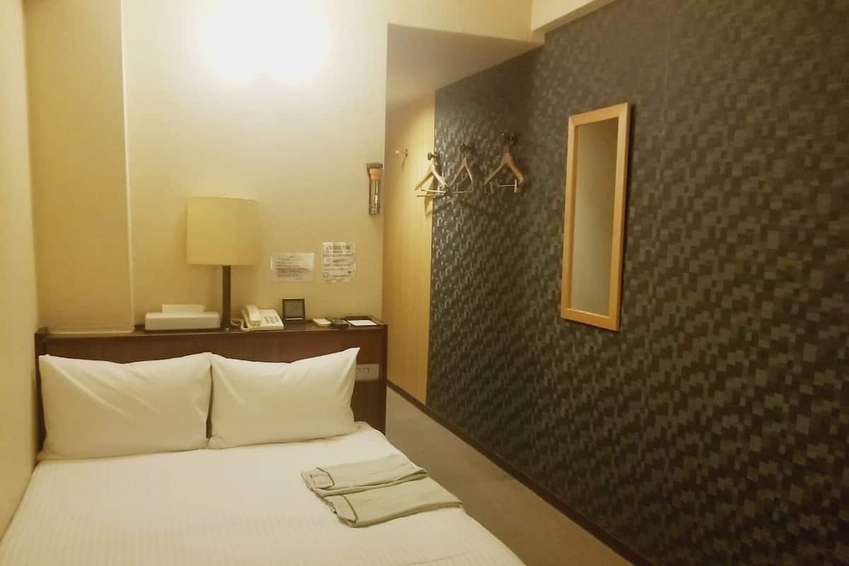 A hotel room with a bed, a desk with a lamp, a telephone, and a mirror on the wall.