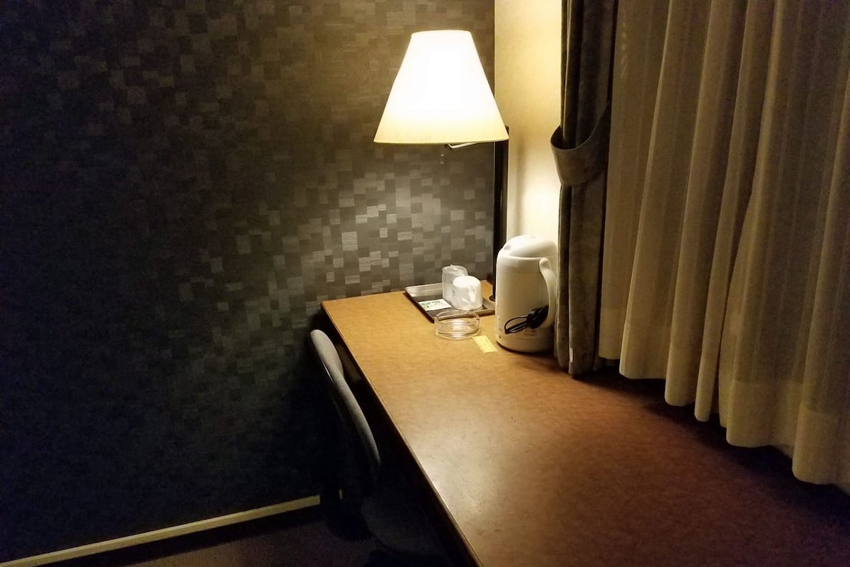A hotel room with a wooden desk, a lamp, a coffee maker, and a curtain.