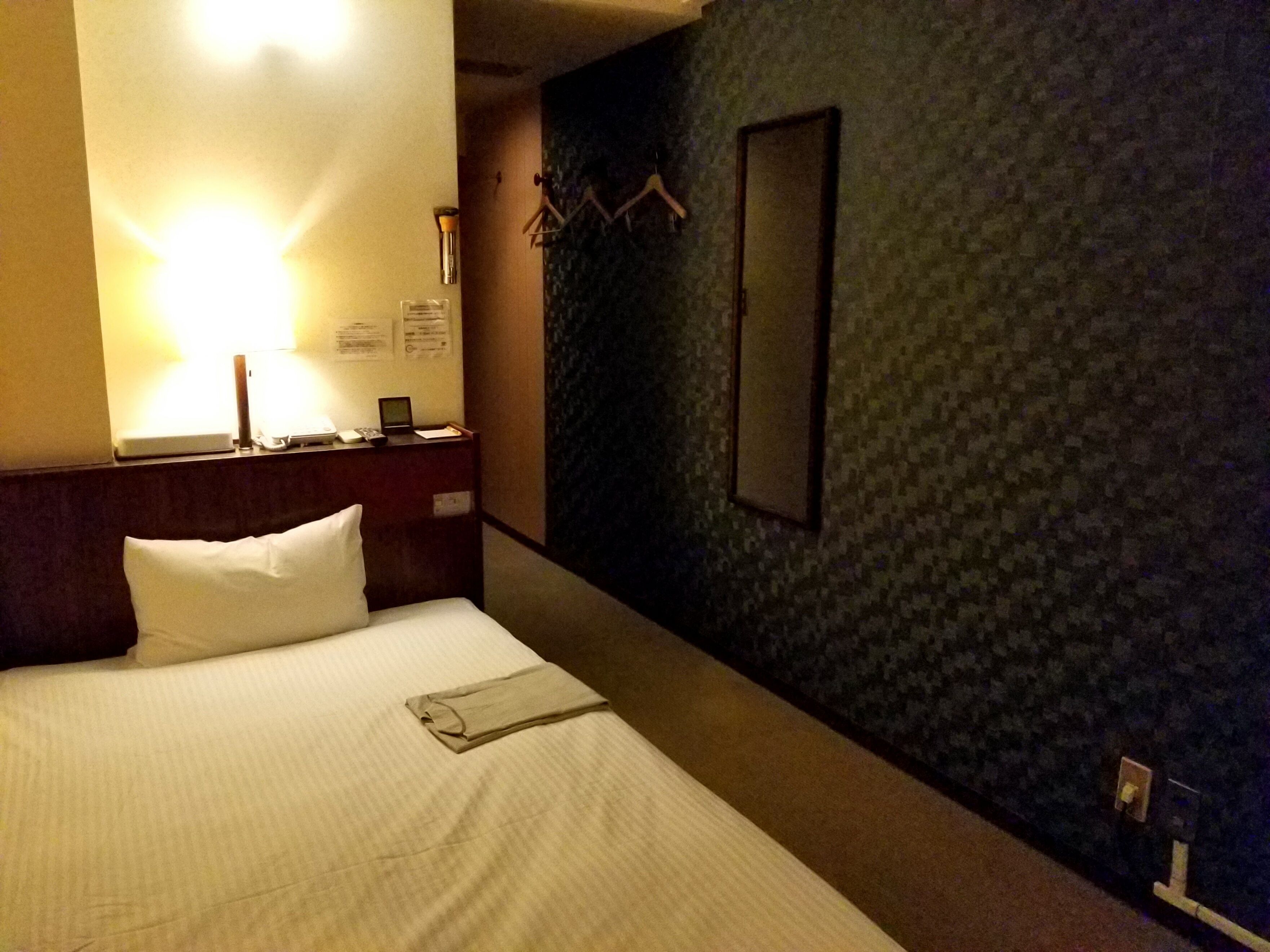 A hotel room with a neatly made bed, a bedside table with a lamp, a mirror, and a wall-mounted coat rack.