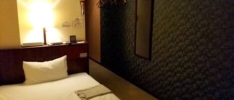 A hotel room with a neatly made bed, a bedside table with a lamp, a mirror, and a wall-mounted coat rack.
