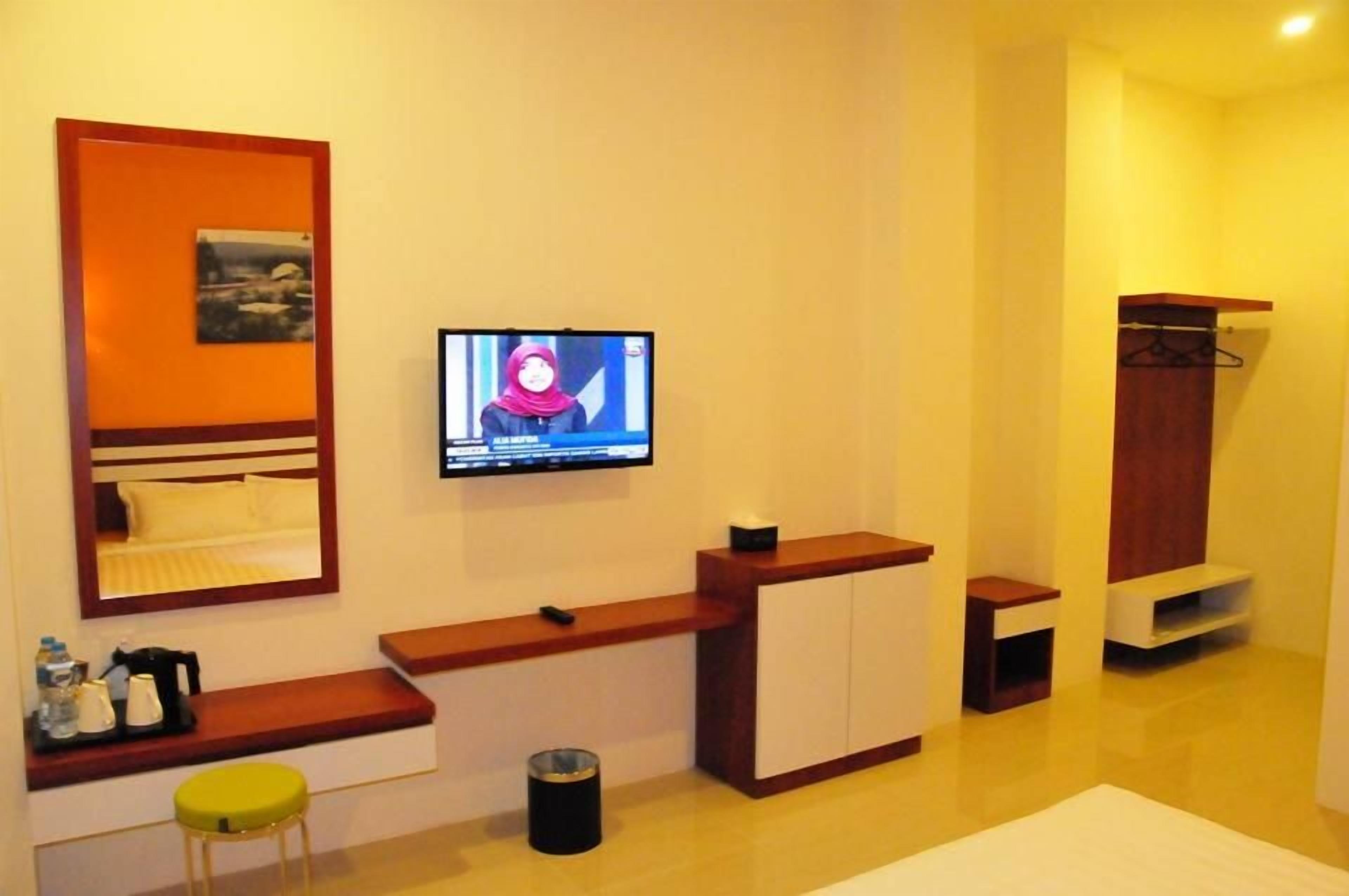 A hotel room with a television mounted on the wall, a bed with a headboard, a small seating area with a stool, a wooden cabinet, and a coat rack.