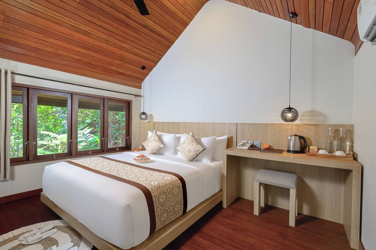 A bedroom with a bed, bedside table, and a view of greenery through a window.