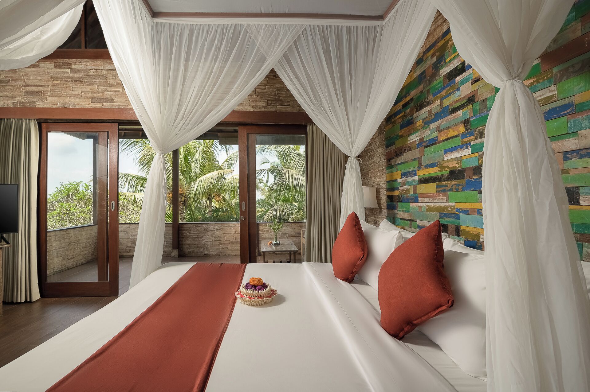 Een slaapkamer met een groot bed, witte gordijnen, een kleurrijke muur en uitzicht op palmbomen.