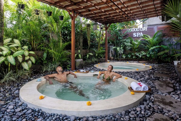 Un espace spa comprenant deux bains à remous entourés de rochers et de plantes tropicales, le tout abrité sous une pergola en bois.