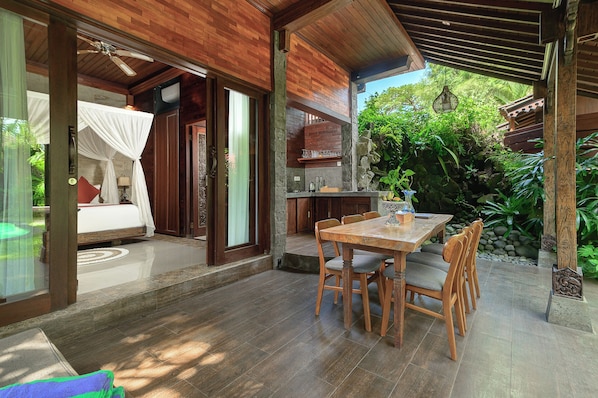 A room with a bed, a dining table with chairs, a kitchen area, and a view of greenery. - Hideaway Villas Candy - Ubud by Kanaan Hospitality (Ubud)