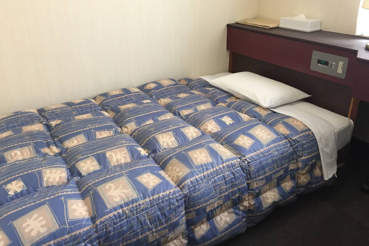 A single bed with a blue and beige patterned duvet, a white pillow, and a small bedside table with a tissue box.